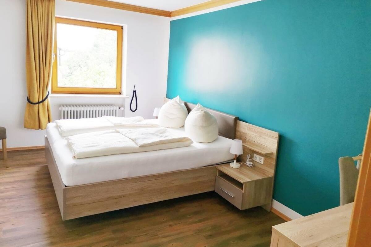 Un dormitorio moderno con una cama de madera, una mesita de noche, un ventanal con cortinas y una pared de acento color turquesa.
