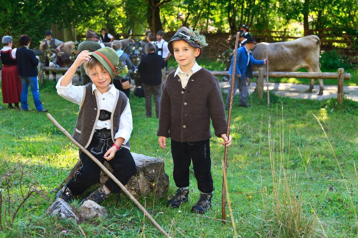 Two children in traditional attire sitting and standing in a grassy outdoor area with a cow in the background.
