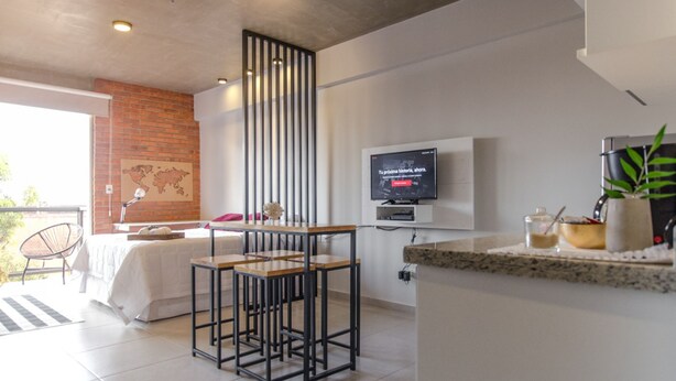 A modern hotel room with a bed, a dining area with stools, a TV mounted on the wall, and a kitchen counter with a plant. - Soho Style Moderno Studio las Mercedes (Asunción)