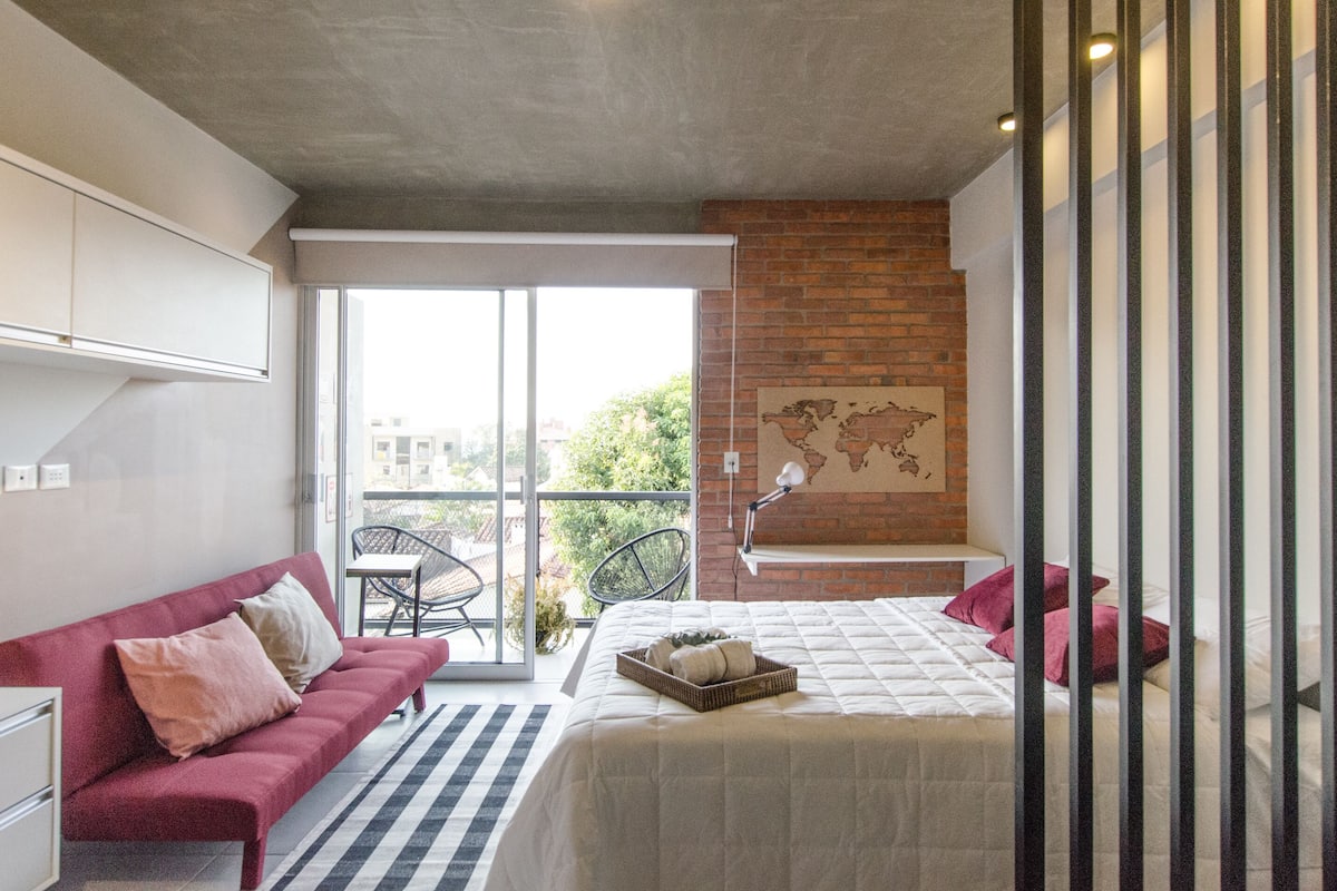 A modern bedroom with a large bed, a sofa, a desk, and a view of the outdoors.