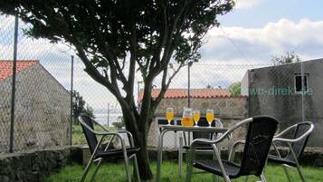 Un espace extérieur aménagé avec des chaises et une table en métal, deux verres de jus d’orange et un arbre qui offre de l’ombre.