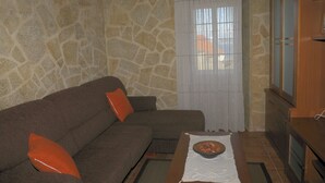 A living room with a stone wall, a brown sofa with orange cushions, a wooden cabinet, and a window with white curtains. - Rustic house with sea views (Carnota)