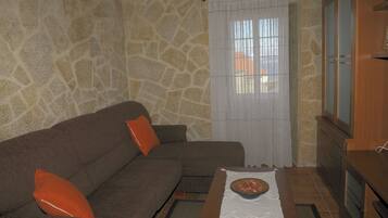Un salon avec un mur en pierre, un canapé marron agrémenté de coussins orange, un meuble en bois et une fenêtre avec des rideaux blancs.