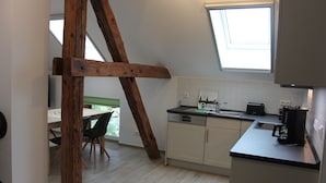 A kitchen with white cabinets, a wooden beam, a window, and a dining area with a table and chairs. - Ferienwohnung Scheune (Lübbenau/Spreewald)