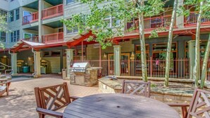 Un espace extérieur aménagé en terrasse, avec des tables et des chaises en bois, un barbecue et une vue sur un immeuble à plusieurs étages doté de balcons.