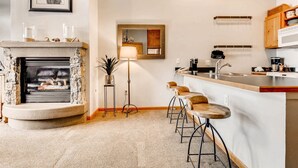 A modern kitchen with a stone fireplace, bar stools, a tall floor lamp, and a wooden cabinet with dishes. - 1 Bedroom Plus Murphy Mountain Condo in River Run Village With Mountain Views and Walking Distance to Ski Slopes (Keystone)