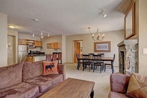 A living room with a brown sofa, a wooden coffee table, and a fireplace. There is a dining area with a table and chairs, and a kitchen in the background. - 3 Bedroom Mountain Condo in River Run Village With Expansive Mountain Views and Walking Distance to Ski Slopes (Keystone)
