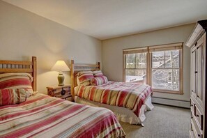 Une chambre avec deux lits, une table de chevet avec une lampe, une fenêtre donnant sur des arbres et une armoire en bois.