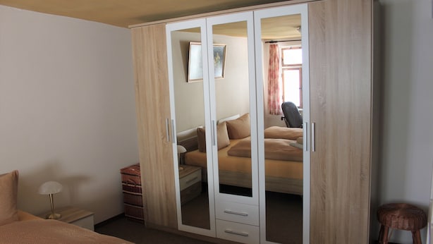 A bedroom with a large wardrobe, a bed, a nightstand, and a wicker stool. - Ferienwohnung Brunnenblick (Lübbenau/Spreewald)