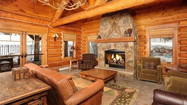 L’intérieur d’une cabane en rondins, avec une cheminée en pierre, des meubles en bois et une grande fenêtre donnant sur un paysage enneigé.