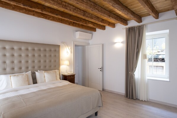 Quarto com uma cama grande, vigas de madeira no teto, mesa de cabeceira com abajur, porta e janela com cortinas.