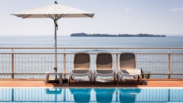 A poolside area with three lounge chairs and a large umbrella, overlooking a calm sea and a distant island. - Hotel Villa Rosenhof 1893 (Gardone Riviera)