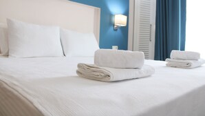 Cama bem arrumada com lençóis brancos, uma luminária fixada na parede e uma janela com cortinas azuis.