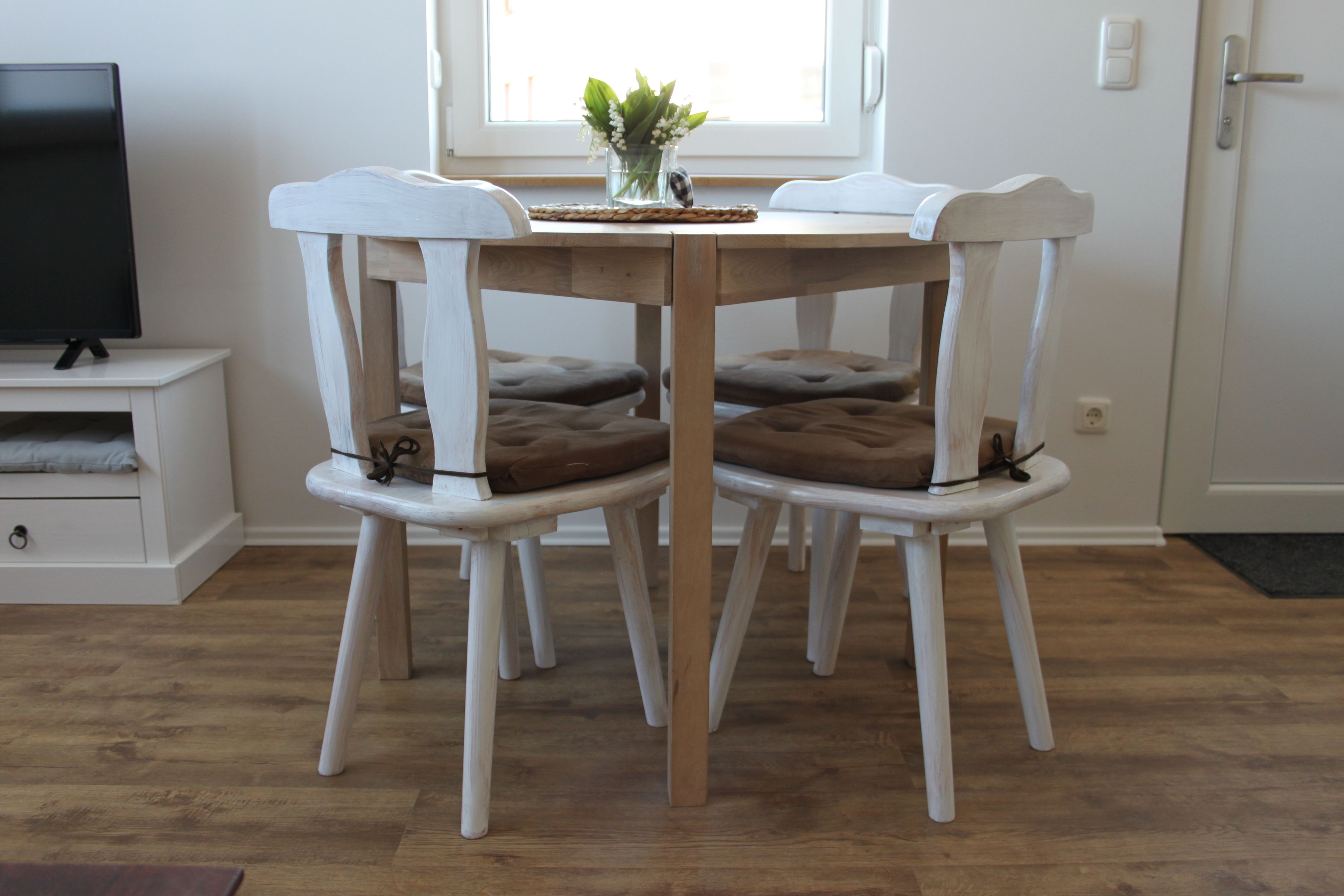 Un coin repas avec une table et des chaises blanches, un vase rempli de fleurs et un téléviseur posé sur un meuble.