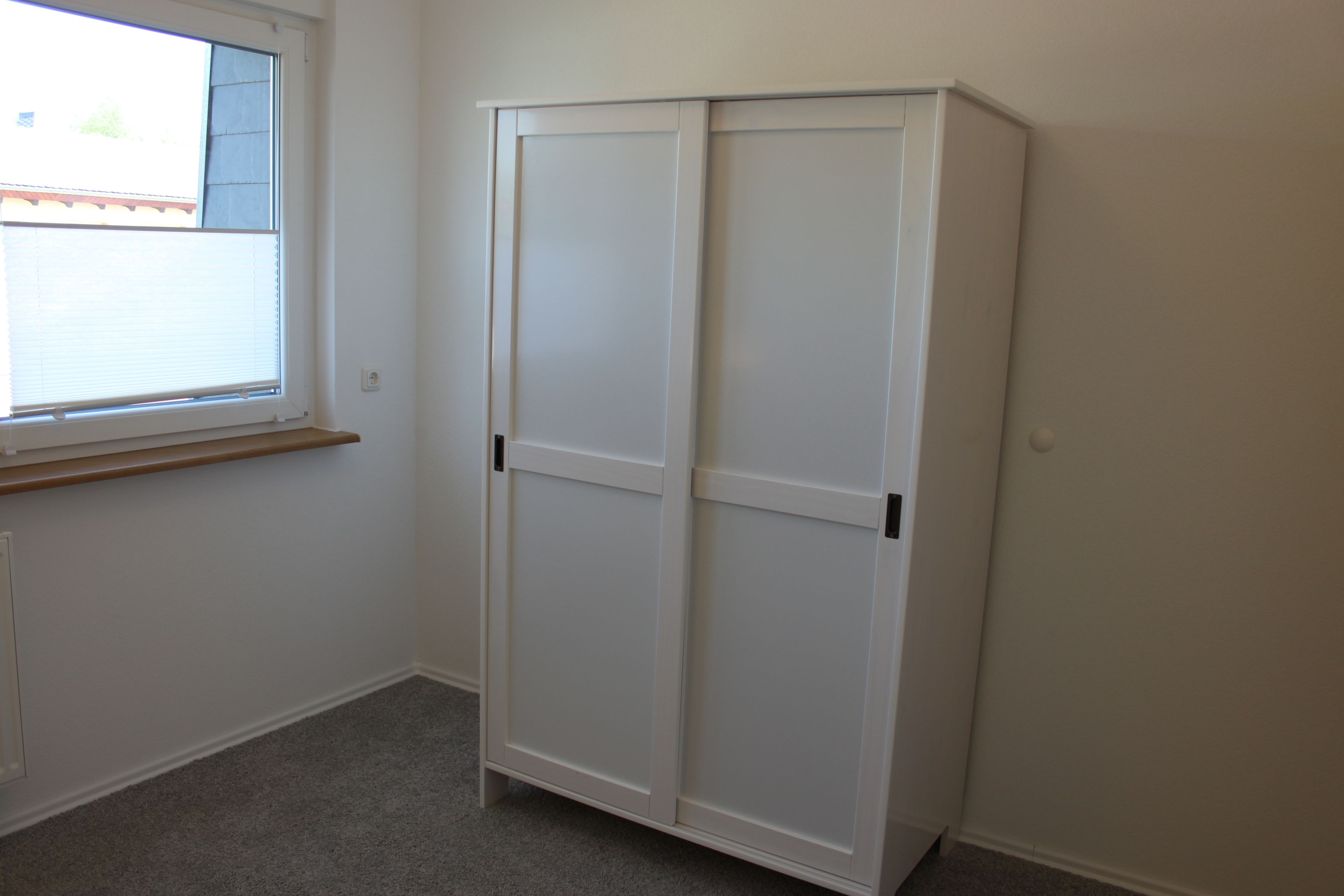 A room with a white wardrobe, a window with blinds, and a carpeted floor.