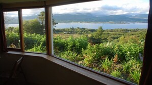 A room with a large window offering a view of a lake, mountains, and greenery. - Beara jewel, Simple but complete appartment style property with stunning view. (Kenmare)