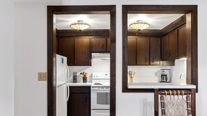 Una cocina con armarios de madera oscura, un refrigerador blanco y una estufa. Hay una mesa de comedor con una silla y una toalla colgada en la silla.