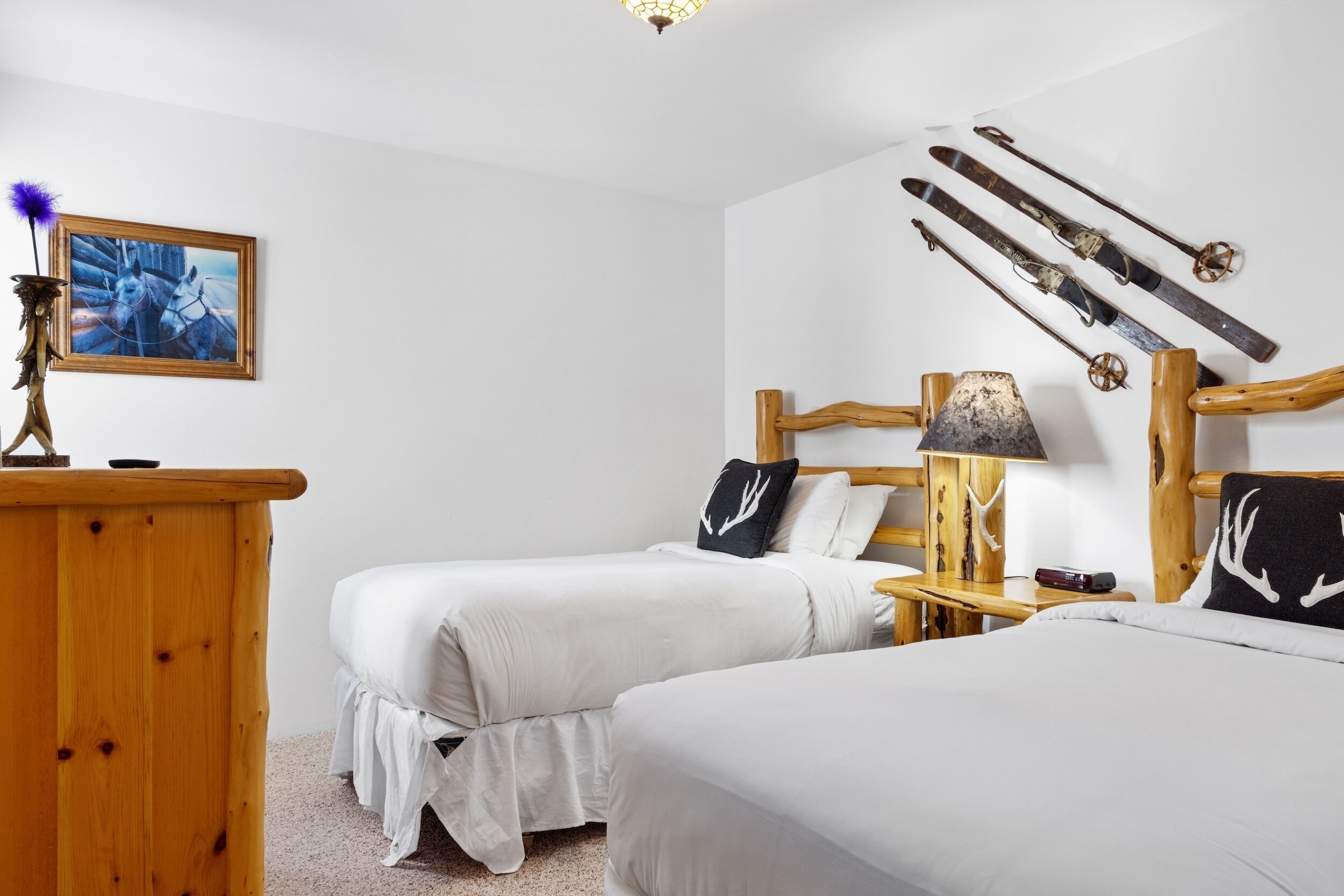 A room with two beds, a wooden dresser, a painting, and skis mounted on the wall.