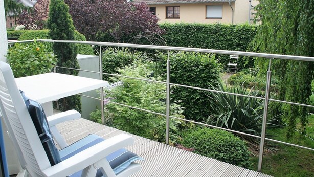 A balcony with a white table and chairs, surrounded by metal railings and lush greenery.