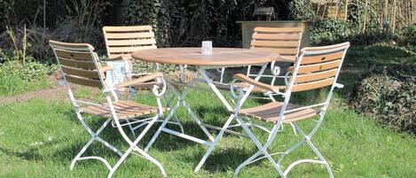 Una mesa redonda de madera con cuatro sillas, rodeada por un jardín con seto y una caseta pequeña al fondo.