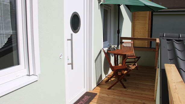 Une terrasse en bois avec une table et des chaises, une porte sur laquelle est posé un paillasson avec le mot « MAISON », et un parasol vert.
