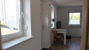 Une pièce avec une fenêtre, une porte, un bureau avec une chaise, un petit tabouret et un téléviseur.
