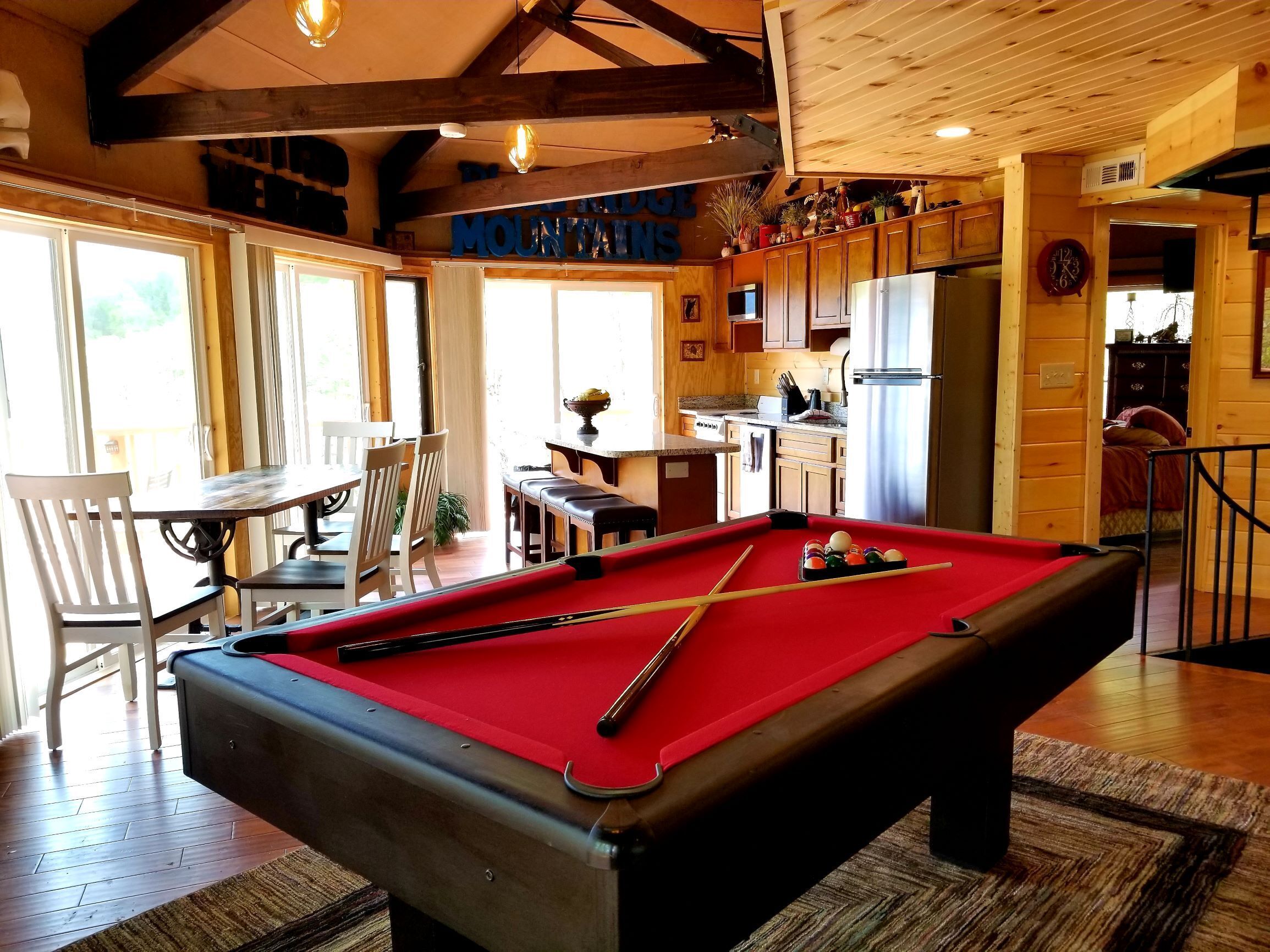 A pool table in a room with wooden beams, a dining area with chairs, and a kitchen area with wooden cabinets.