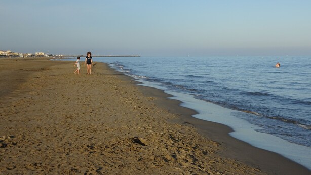 Een strand met een heldere lucht, een pier in de verte en enkele mensen vlakbij het water.