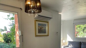 Een eethoek met een ronde tafel, stoelen, een vaas met bloemen, een hanglamp, een airconditioner, een lijst met een afbeelding en een bank op de achtergrond.