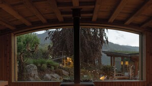 A wooden deck with a central wood-burning stove, a large window overlooking a mountainous landscape, and a cozy seating area. - Naturhotel Lüsnerhof (Luson)