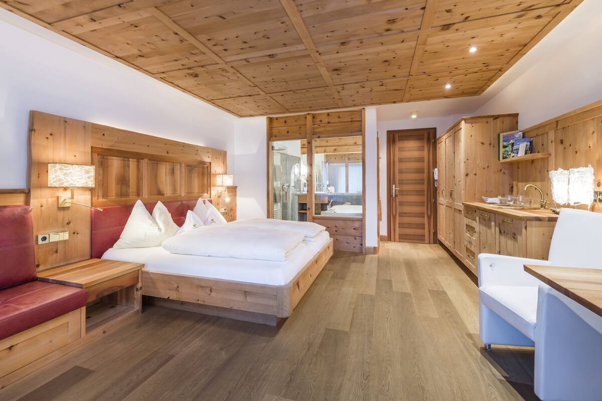 Une pièce aménagée comme un chalet en bois, avec un lit, un banc recouvert d’un tissu rouge, un bureau en bois et une salle de bain visible à travers une porte en verre.
