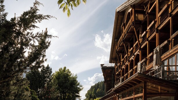 A woman sitting by a pool with a stone patio, surrounded by lush greenery and a wooden building with balconies. - Naturhotel Lüsnerhof (Luson)