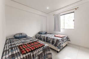 A room with two beds, each with a plaid bedspread and a blue pillow. There is a window with a curtain, and the room has white walls and a tiled floor.
