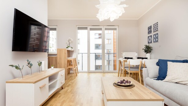 A modern living room with a flat-screen TV, wooden furniture, a dining area, and a large window with a city view. - Business Mokotow Park 214 Apartment (Warsaw)