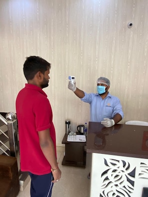 A hotel reception area with a reception desk, a guest standing, and a staff member holding a thermometer. - Hotel Gopal (Haridwar)