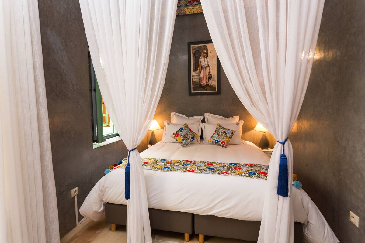 A bed with white bedding, a colorful floral bedspread, and white curtains.