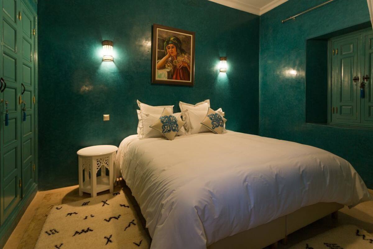 A bedroom with teal walls, a bed with white bedding, a small round table, and a painting above the bed.