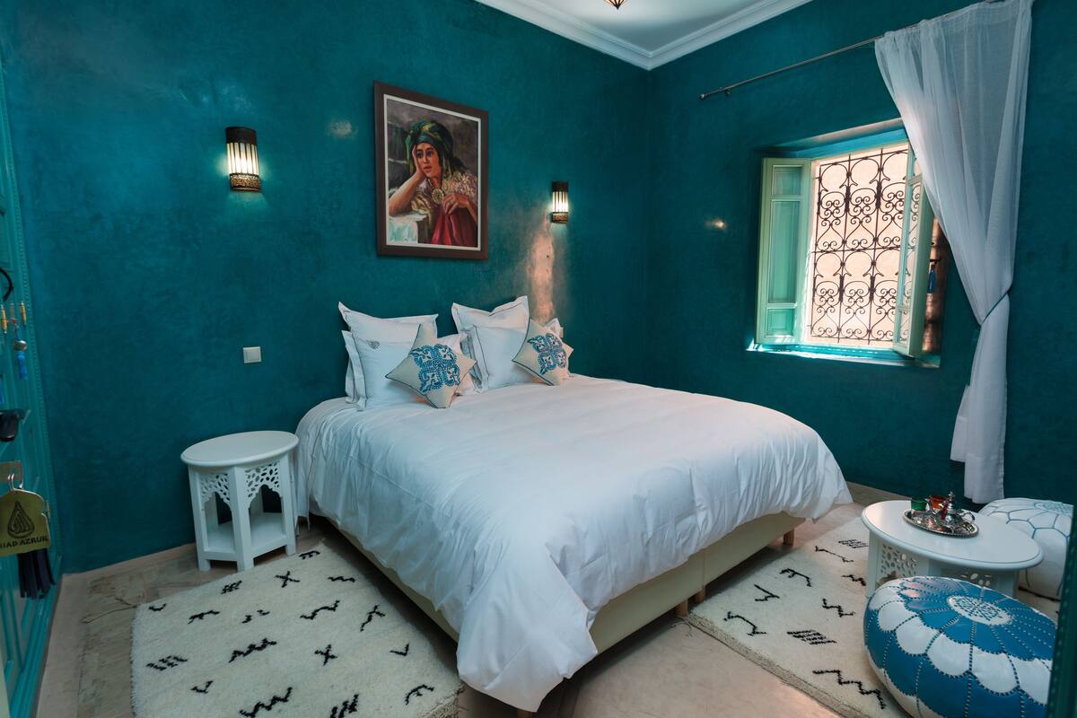 A bedroom with teal walls, a bed with white bedding, a small round table, and a window with a decorative screen.