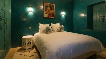 A bedroom with teal walls, a bed with white bedding and patterned pillows, a small white side table, and a painting above the bed.