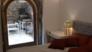 Une chambre avec un lit, un bureau et une lampe. On y trouve un mur en pierre et une porte qui donne sur un espace extérieur aménagé pour se détendre.