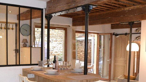 Un espace repas avec une table en bois et des chaises blanches, un mur en pierre et un plafond en bois.
