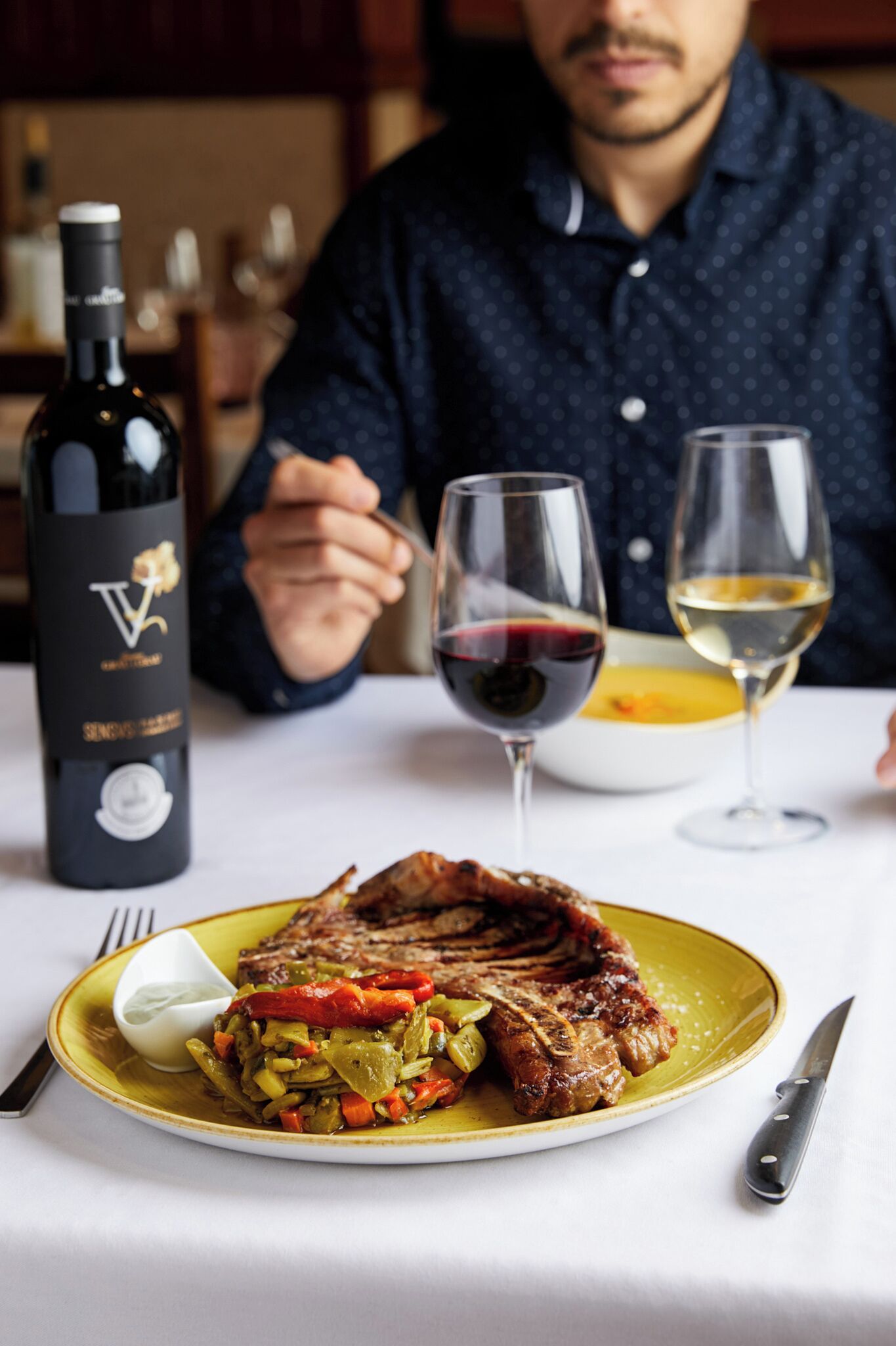 Een eettafel met een steak, groenten, een fles wijn en twee wijnglazen.
