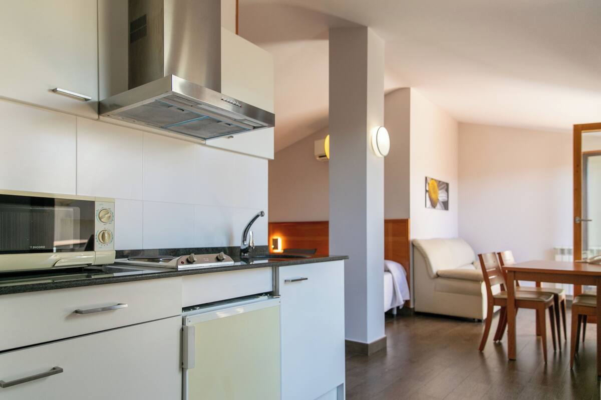 A modern kitchen with a built-in microwave, stove, and ventilation hood. There is a dining area with a table and chairs, and a sofa in the background.