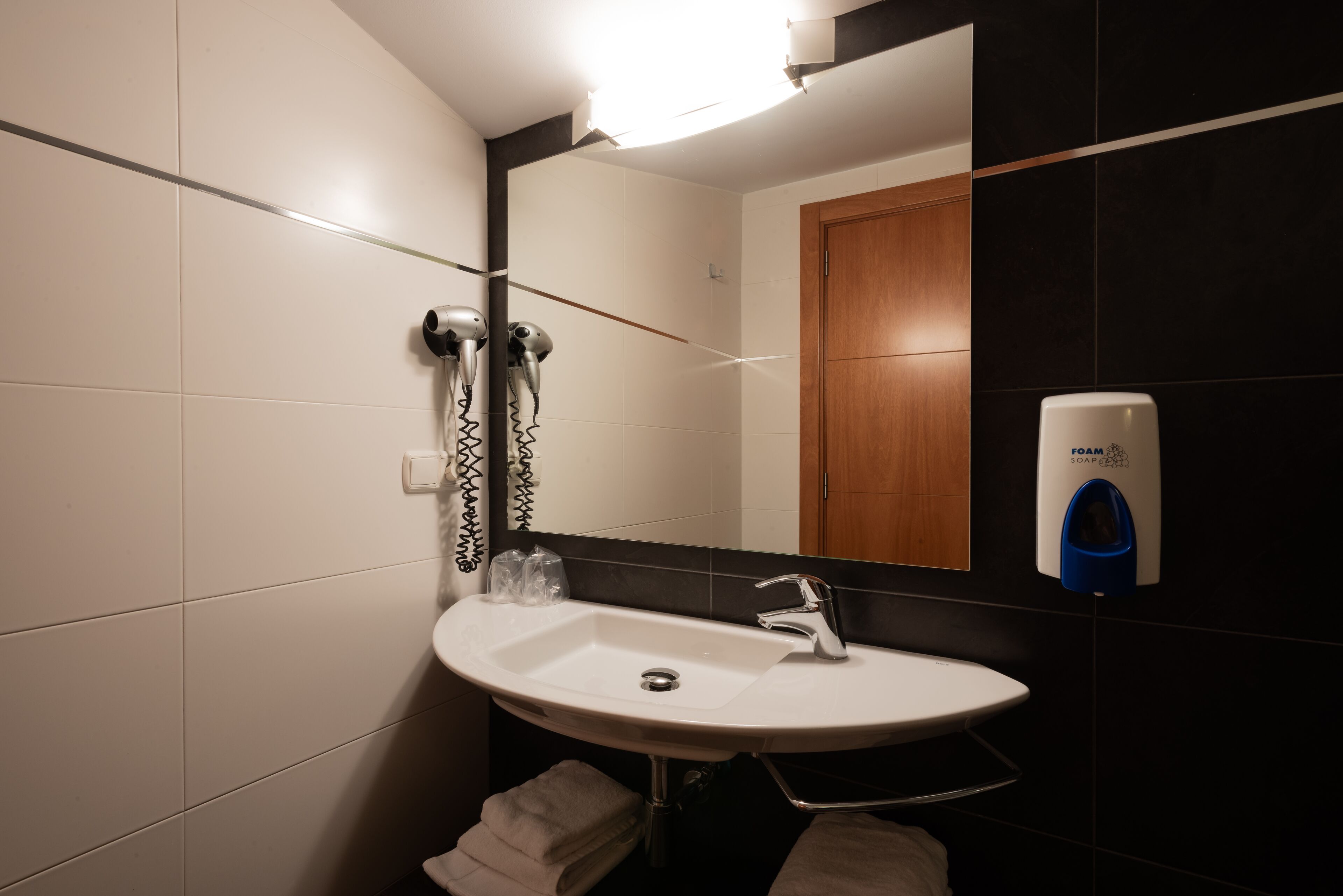 Un baño con lavamanos blanco, un espejo, un secador de pelo y un dispensador de jabón.