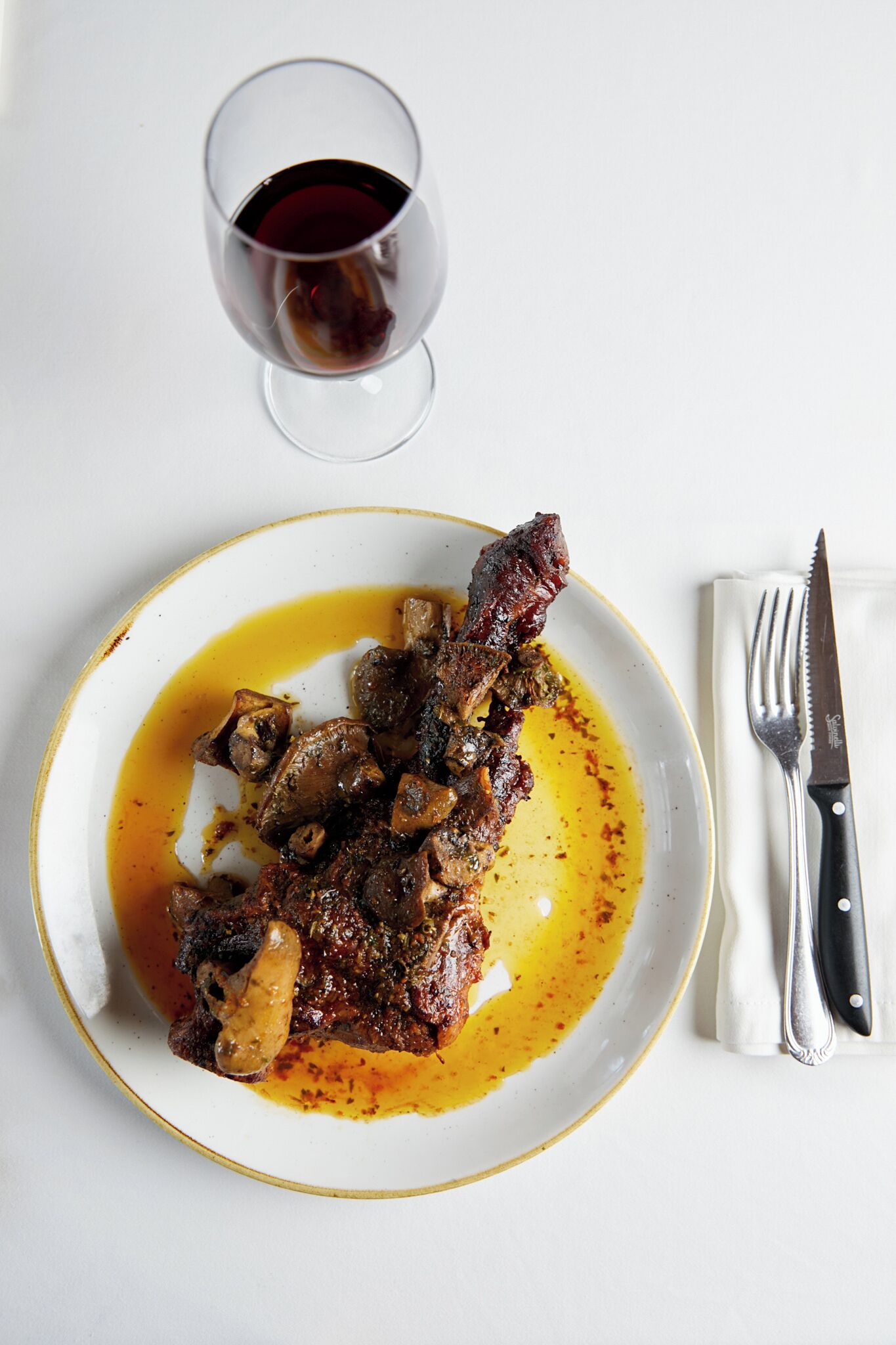 A plate of meat with sauce and a glass of red wine on a white table.