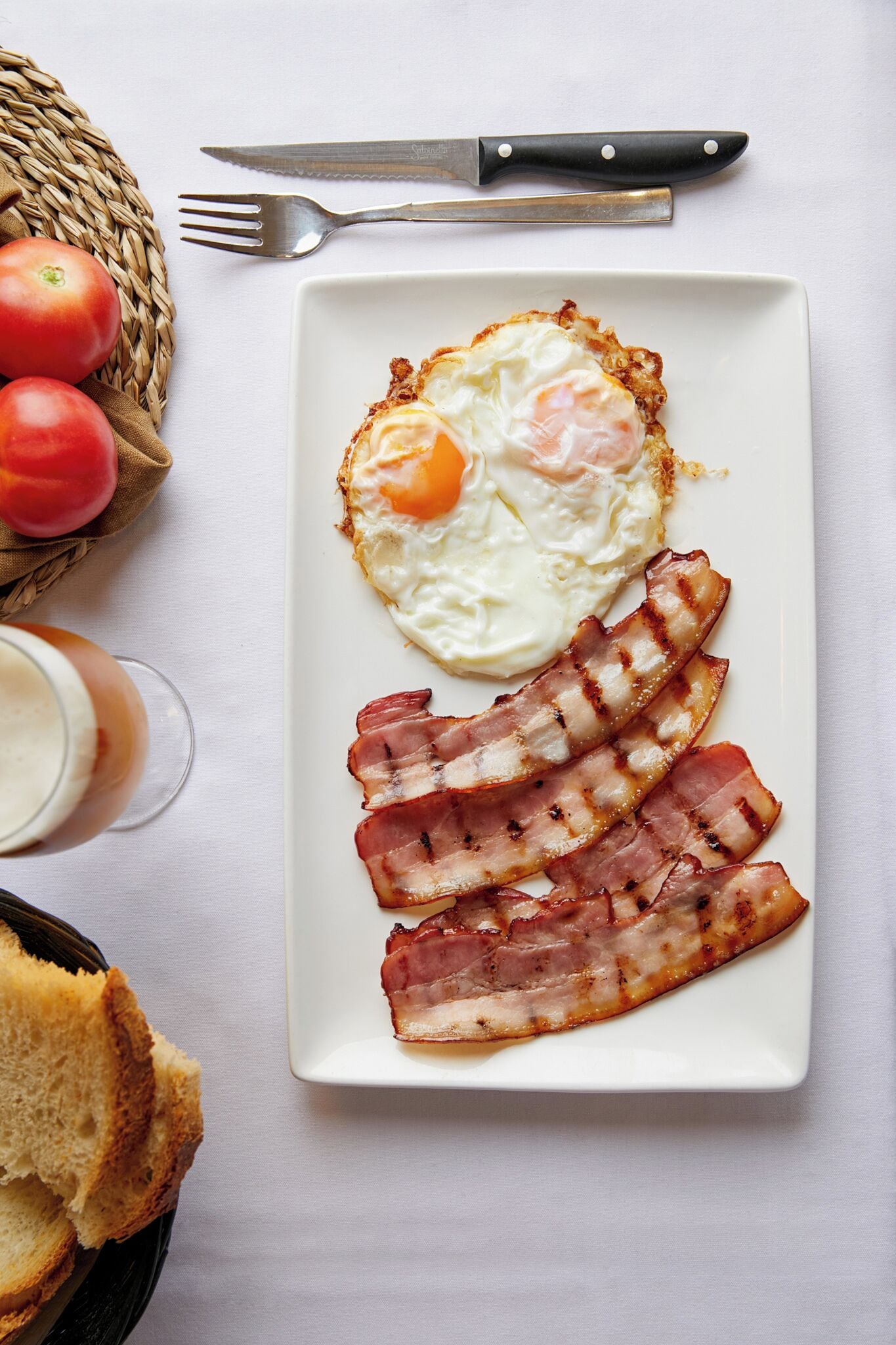 Un plato de desayuno con huevos y tocino, un tenedor, un cuchillo y un vaso de bebida sobre una mesa.
