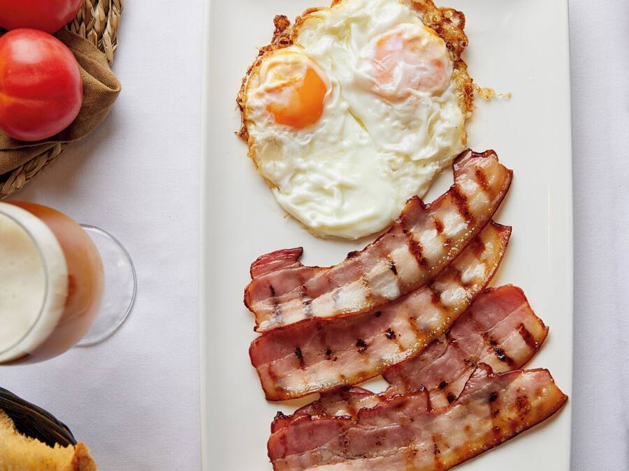 Un plato de desayuno con huevos y tocino, un tenedor, un cuchillo y un vaso de bebida sobre una mesa.