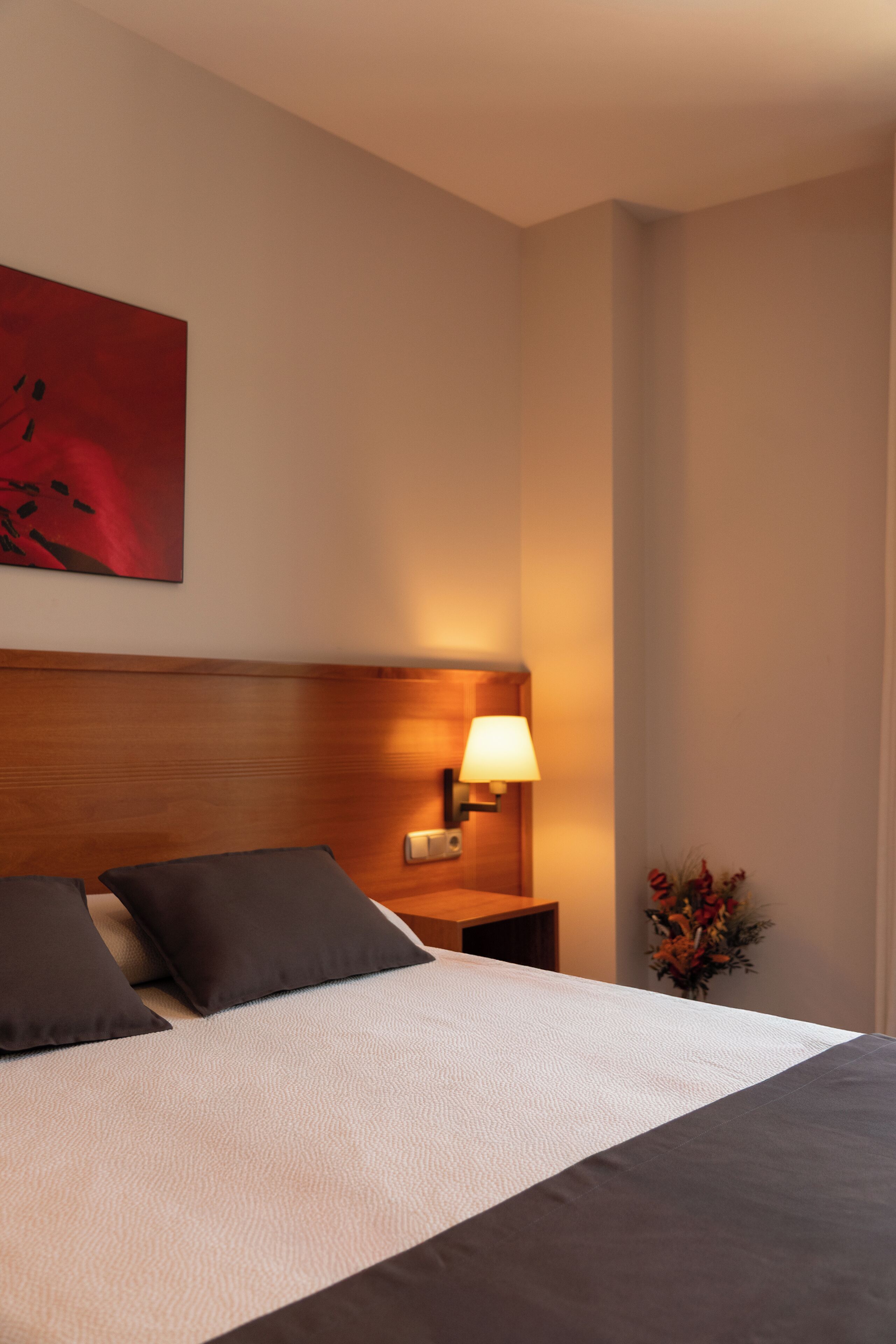 Una cama bien hecha con almohadas oscuras y una colcha blanca, una lámpara de noche y un cuadro abstracto rojo en la pared.