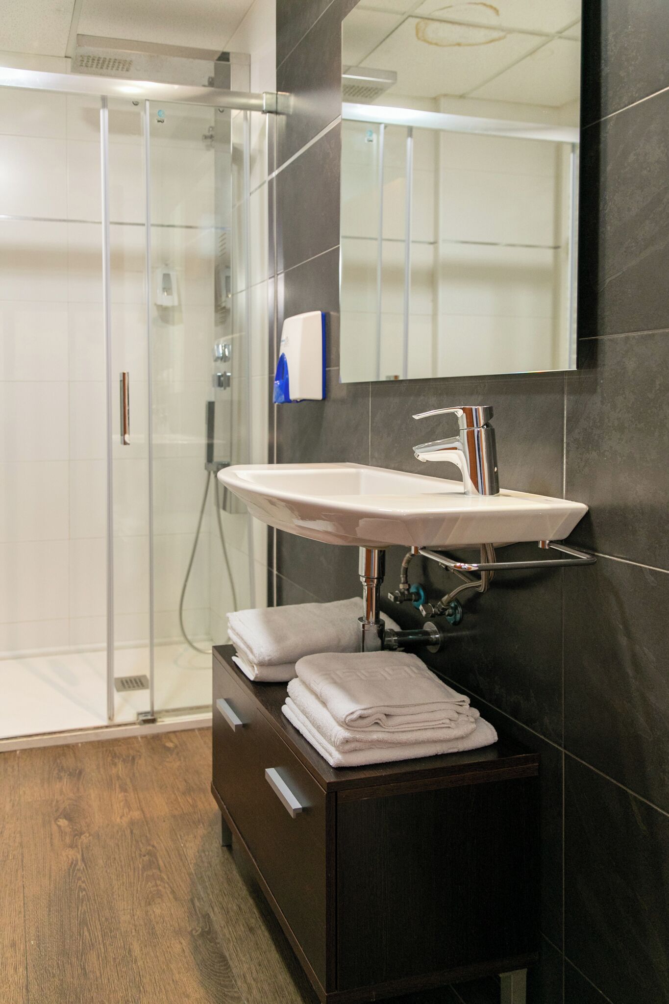A modern bathroom with a glass shower enclosure, a white sink with chrome fixtures, and dark tiled walls.