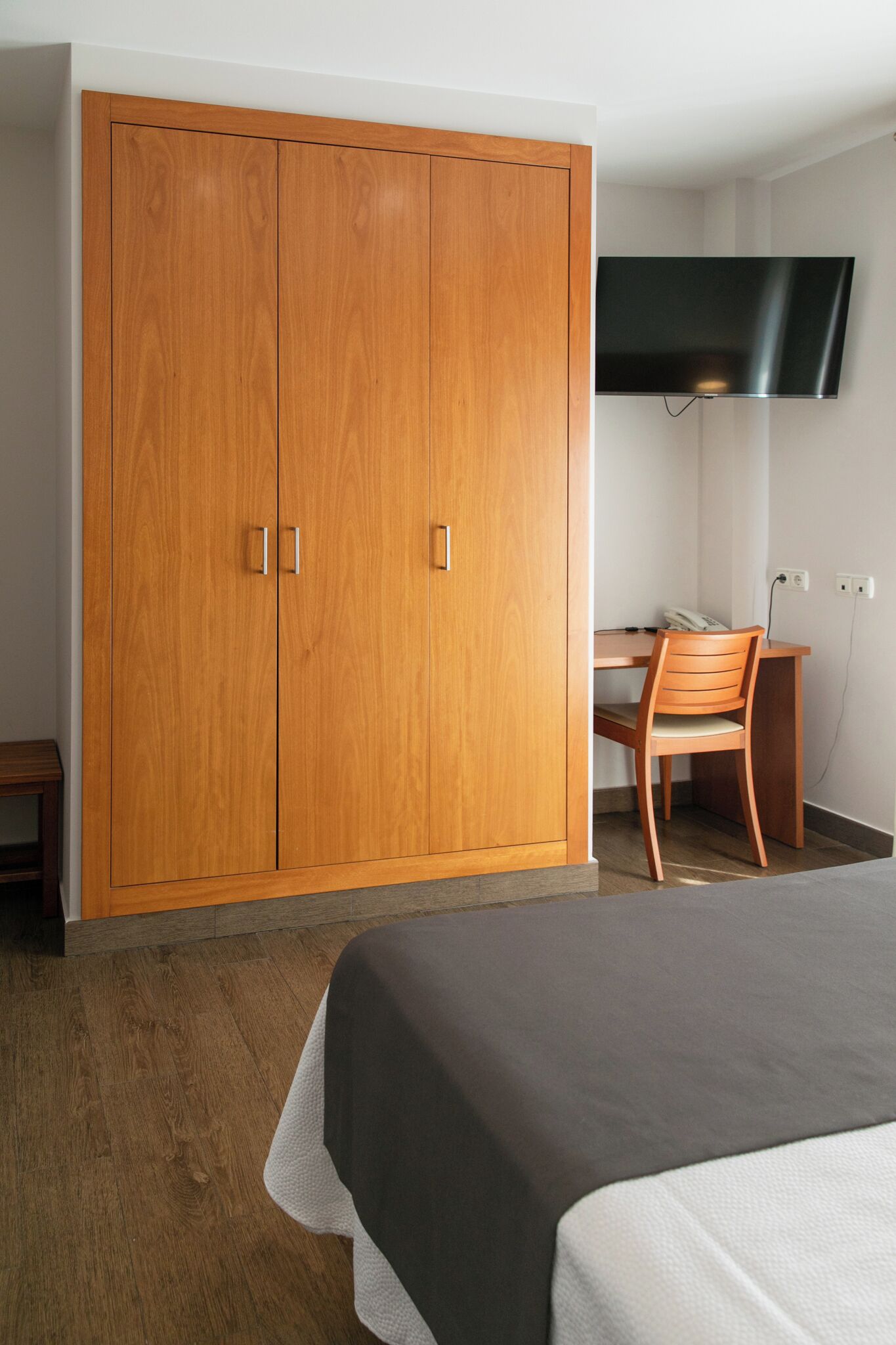 A bedroom with a wooden wardrobe, a desk with a chair, a bed, and a TV mounted on the wall.